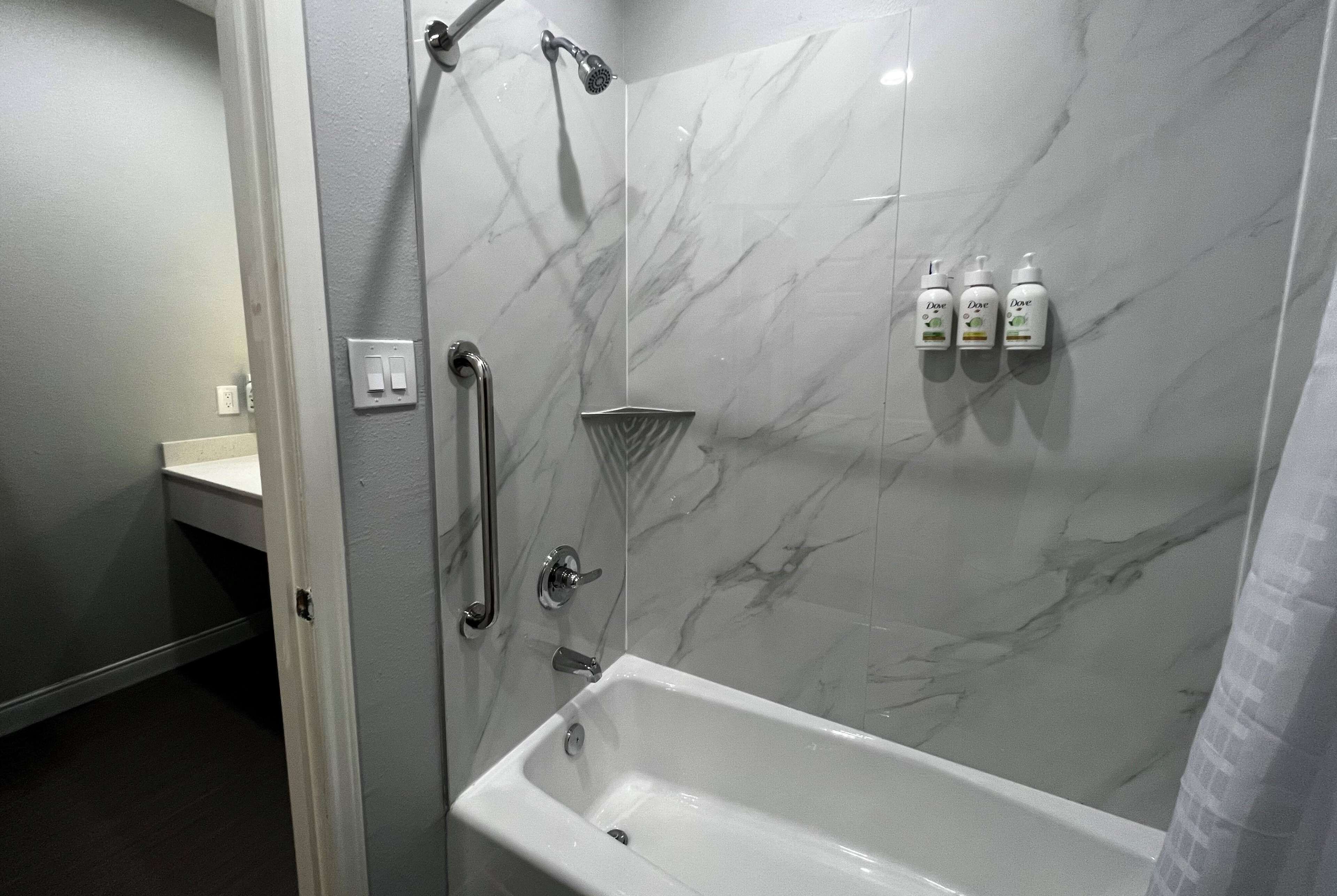 Un baño con pared de ducha de mármol, una bañera blanca y tres botellas de Dove para el cuerpo en la pared.