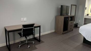 A hotel room with a wooden desk, a black office chair, a television on a wooden cabinet, a bed with white bedding, and a small trash bin.