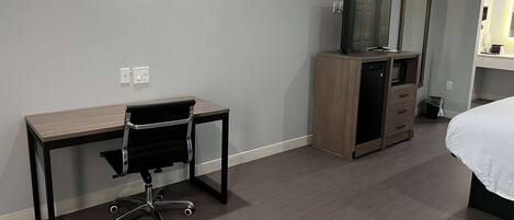 Habitación de hotel con un escritorio de madera, una silla de oficina negra, un televisor sobre un mueble de madera, una cama con ropa blanca y un cubo de basura pequeño.