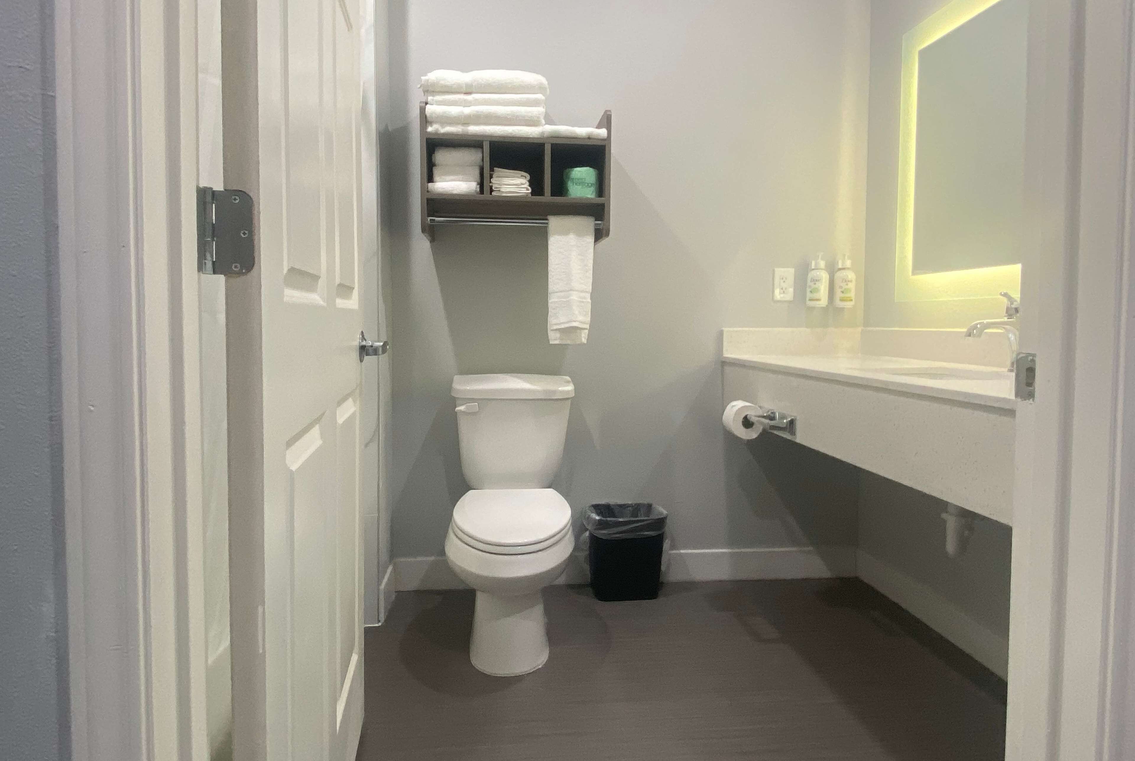A compact bathroom with a toilet, a shelf with towels, a trash bin, and a sink with a mirror.