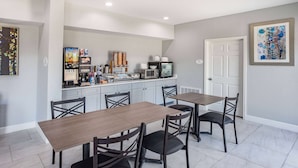 A dining area with a long wooden table and four chairs, a kitchen area with appliances and a refrigerator, and a door with a framed picture on the wall.