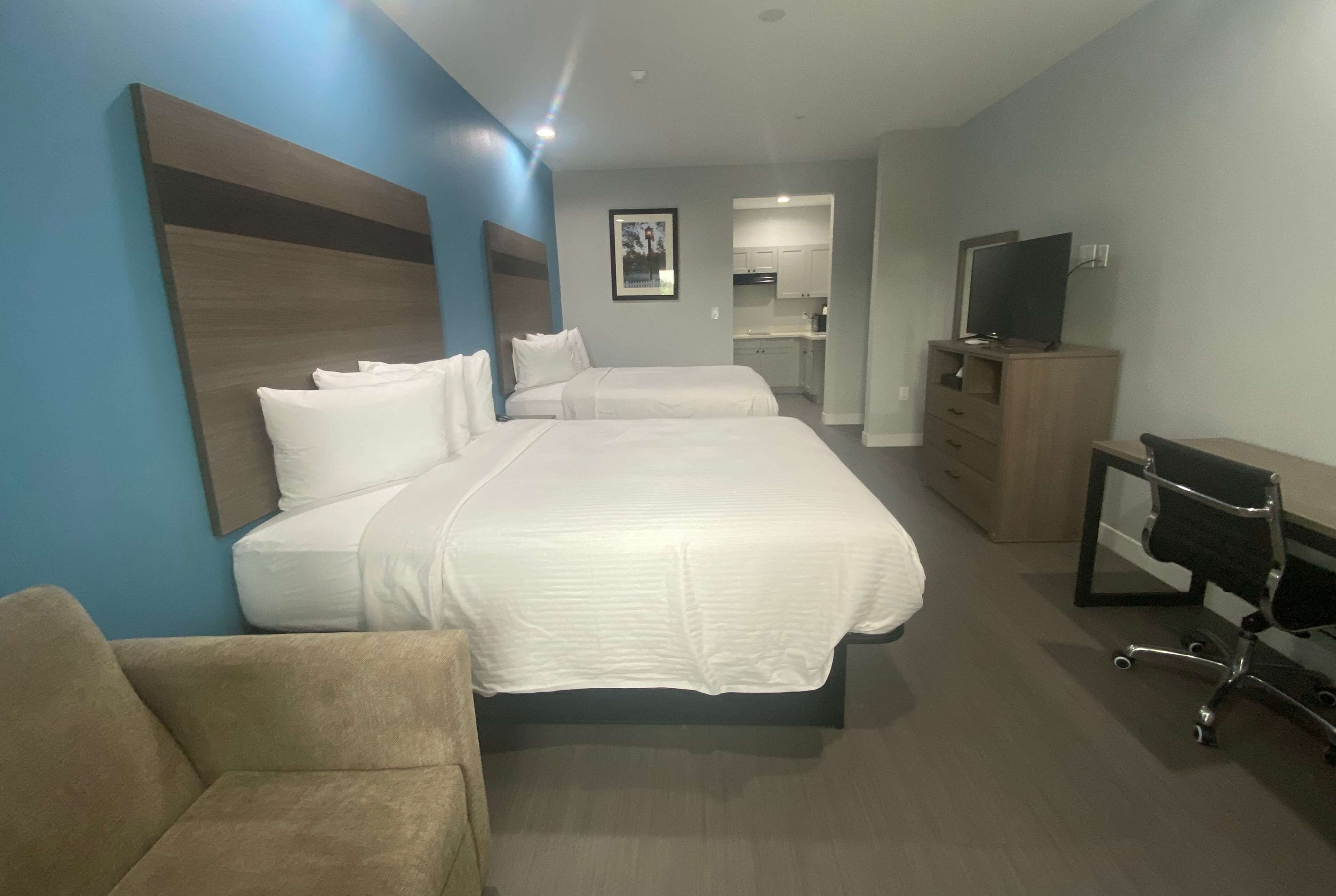 A hotel room with a large bed, a sofa, a TV, and a kitchenette in the background.