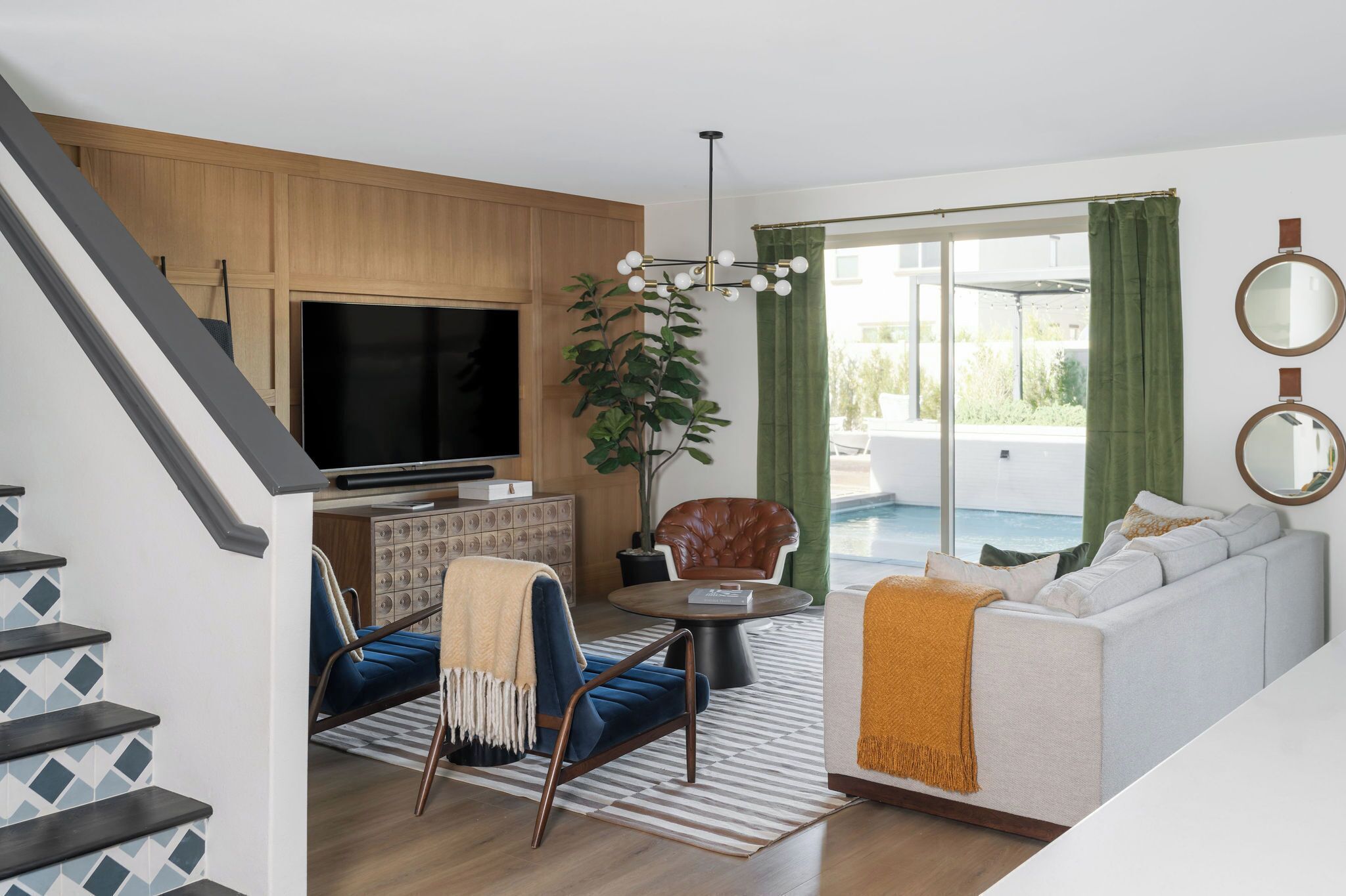 Una sala de estar moderna con televisor, sofá y sillones. Se observa una escalera a la izquierda y un balcón con cortinas.