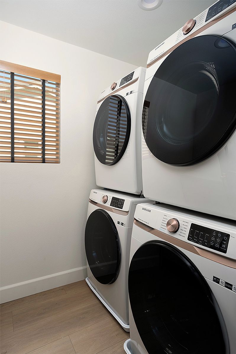 A laundry room with stacked front-loading washing machines and a window with blinds.