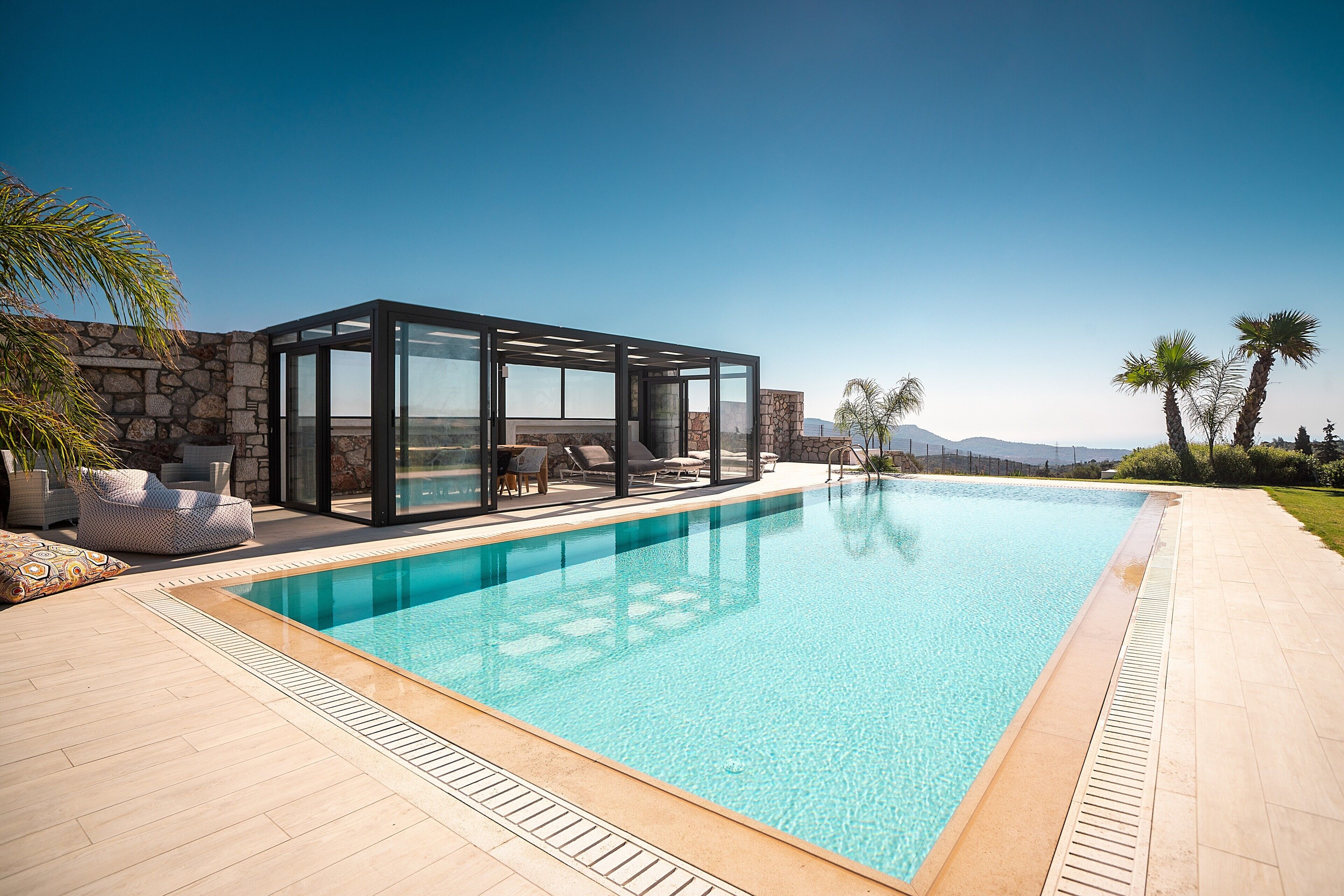 A modern pool area with a glass-enclosed lounge, stone walls, and a clear view of the mountains.
