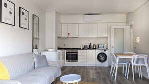 Una cucina moderna con mobili bianchi, forno integrato, microonde e lavatrice. C'è un divano grigio con un cuscino giallo, un tavolino rotondo e una zona pranzo con sedie bianche.