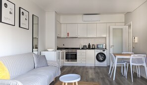 A modern kitchen with white cabinets, a built-in oven, a microwave, and a washing machine. There is a grey sofa with a yellow pillow, a small round table, and a dining area with white chairs.