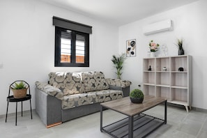A modern living room with a grey sofa, a wooden coffee table, a black chair, a potted plant, a wooden shelf with decorative items, and a window with blinds.