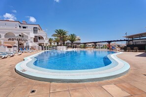 Une piscine avec des chaises longues, un espace de détente couvert et un bâtiment doté de balcons.