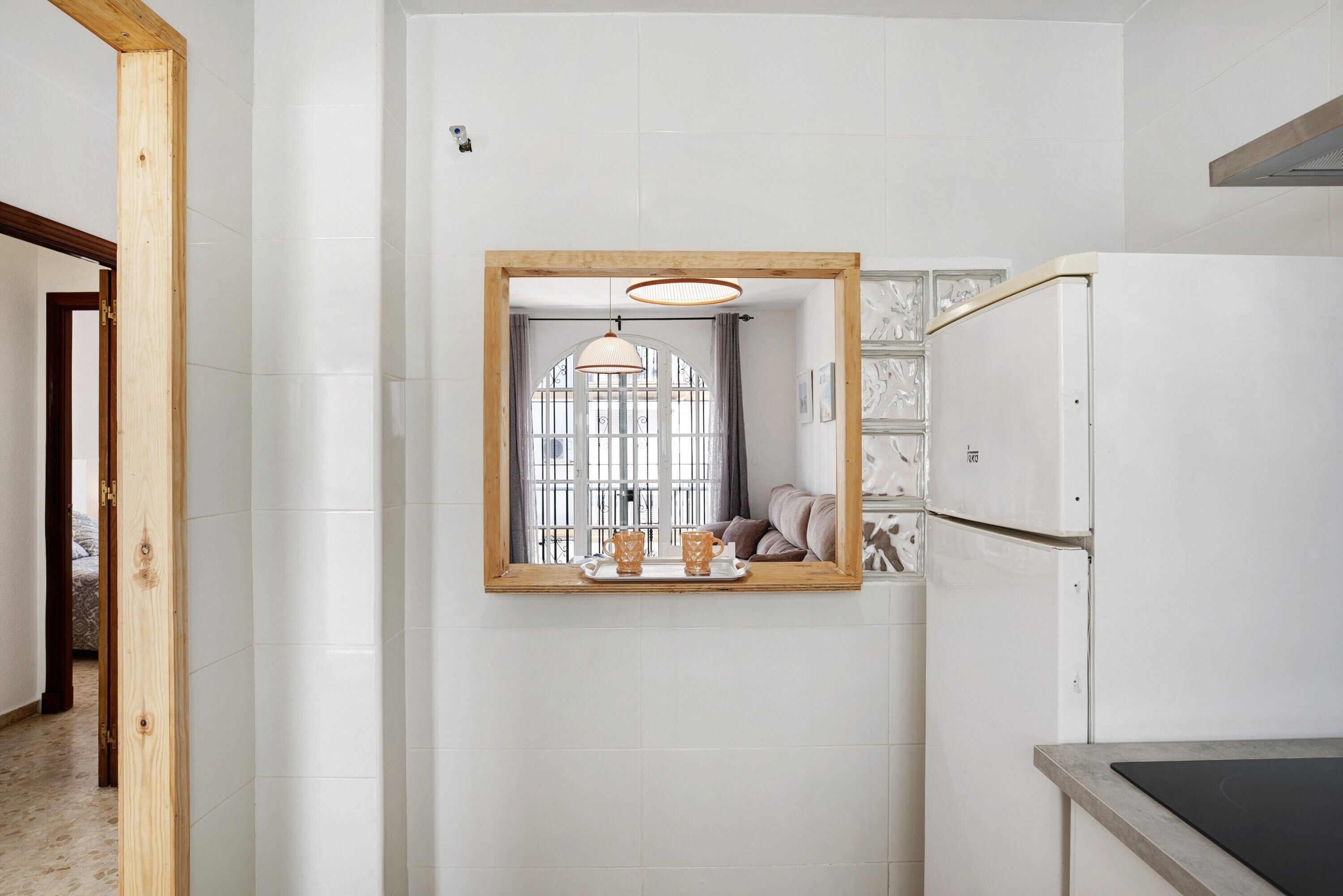 Een keuken met witte betegelde muren, een spiegel met houten lijst, een hanglamp en een koelkast.