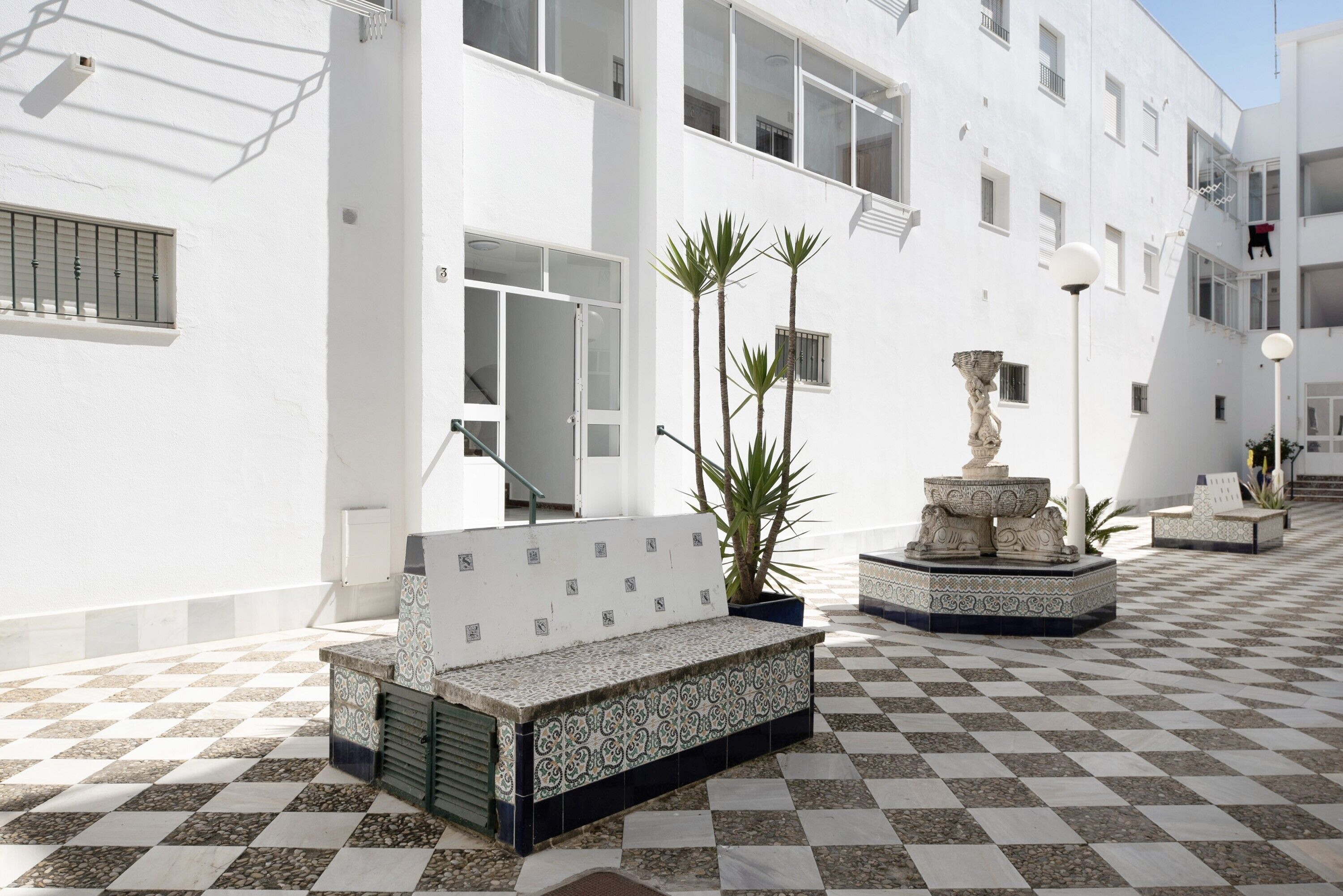 A courtyard with a checkered floor, a bench, a fountain, and a white building with windows and balconies.