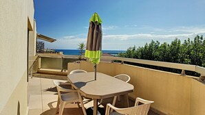 Un balcón con mesa y sillas, una sombrilla verde y vista al mar.