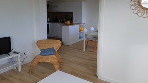 Ein modernes Wohnzimmer mit Fernseher, einem Korbstuhl, einem weißen Tisch und einer Küche im Hintergrund.