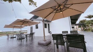 An outdoor patio area with tables and chairs under large umbrellas, a white building in the background, and a view of the landscape. - House in field (Dongshan)