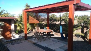 Un patio al aire libre con mesa y sillas de madera, una estructura de techo de paja y vegetación circundante.
