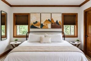 Une chambre à coucher comprenant un lit, deux tables de chevet, une fenêtre avec des stores et trois tableaux accrochés au mur.