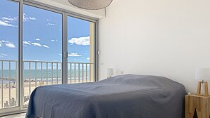 A bedroom with a large bed, a nightstand, a lamp, and a view of the beach and ocean through a large window.