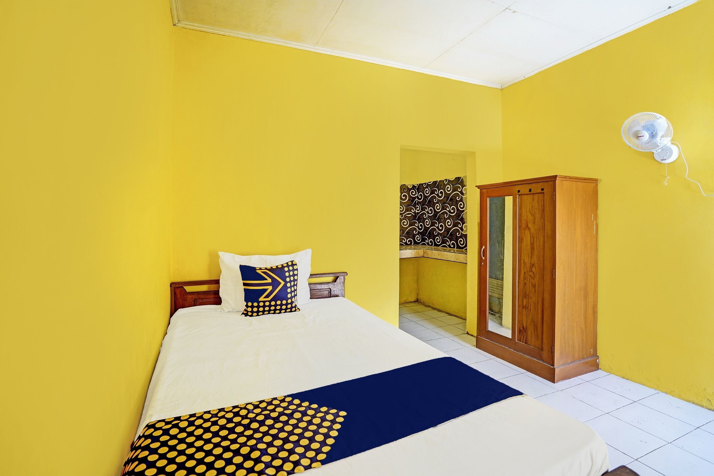 A room with yellow walls, a bed with a white and blue bedspread, a wooden wardrobe, and a white ceiling fan.