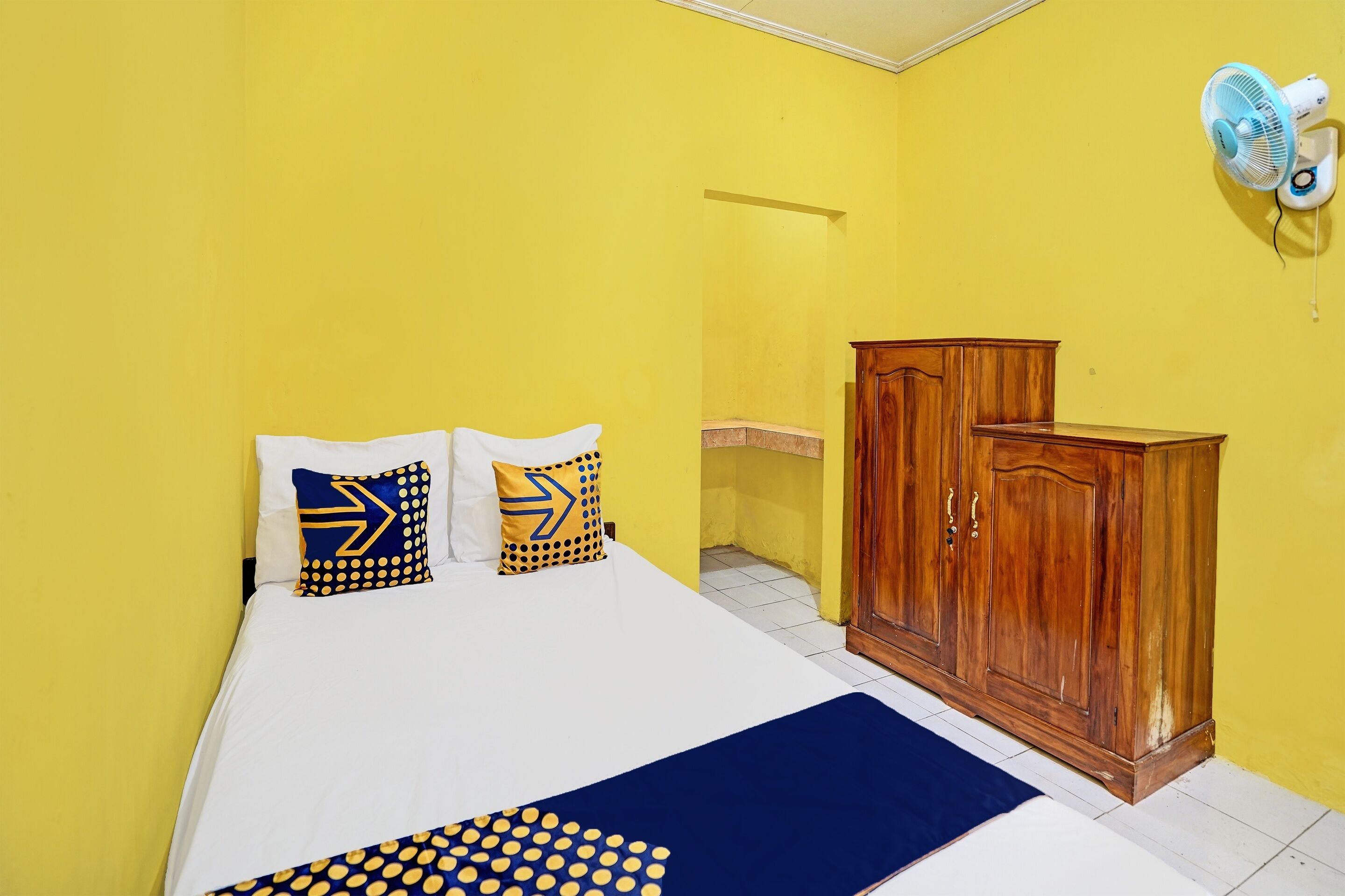 A room with a bed, a wooden wardrobe, and a wall-mounted fan.