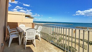 Un balcón con mesa y sillas, con vista a una playa y un cielo despejado.