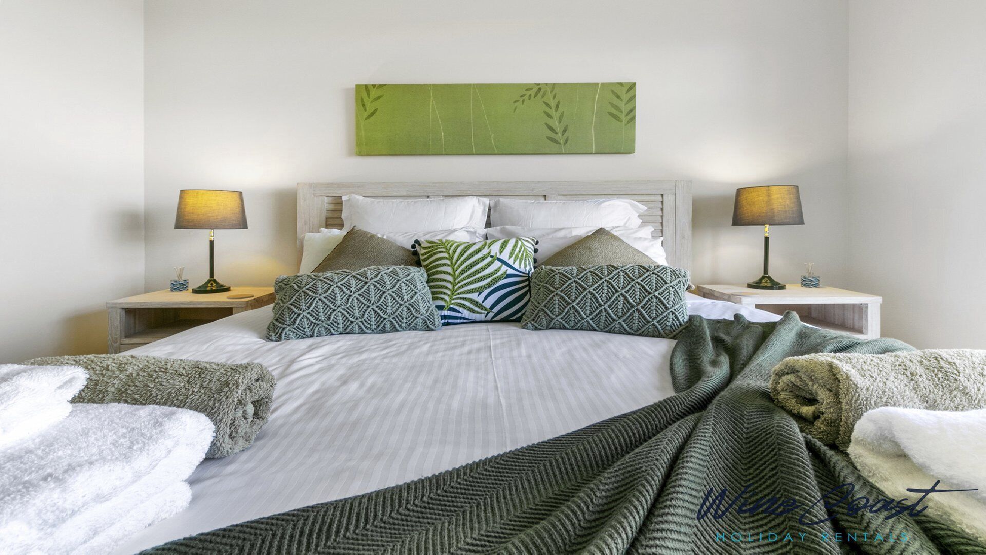 Een net opgemaakt bed met gekleurde kussens en een wanddecoratie met groene bladeren erboven.