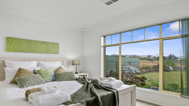 A bedroom with a large window, a bed with multiple pillows, a bedside table with a lamp, and a green wall art piece. - Sellicks Seascape by Wine Coast Holiday Rentals (SELLICKS BEACH)