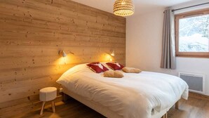 Une chambre avec une tête de lit en bois, un lit avec une literie blanche et des oreillers rouges, un petit tabouret rond et un luminaire suspendu.