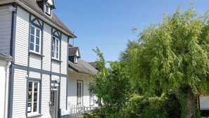 Een huis met twee verdiepingen, een witte gevel en donkere accenten, een houten terras en een grote boom ervoor.