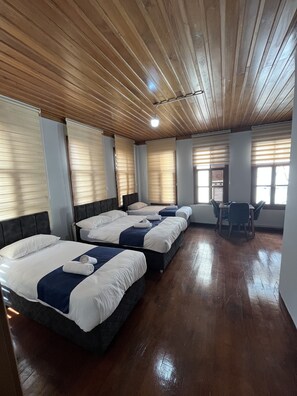 A hotel room with four beds arranged in a row, wooden ceiling, and a dining area with chairs and a table.
