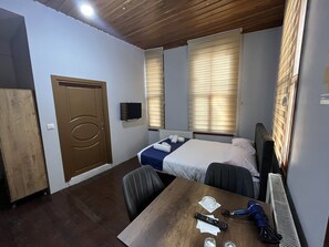 Quarto de hotel com porta de madeira, cama com manta azul, escrivaninha com controle remoto e televisão fixada na parede.