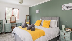 A bedroom with a bed, bedside tables, a dresser, a mirror, and a window with blinds. - Mill End (Penrith)
