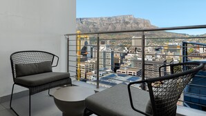 A balcony with two wicker chairs and a small table, overlooking a cityscape with buildings and a prominent mountain in the background.