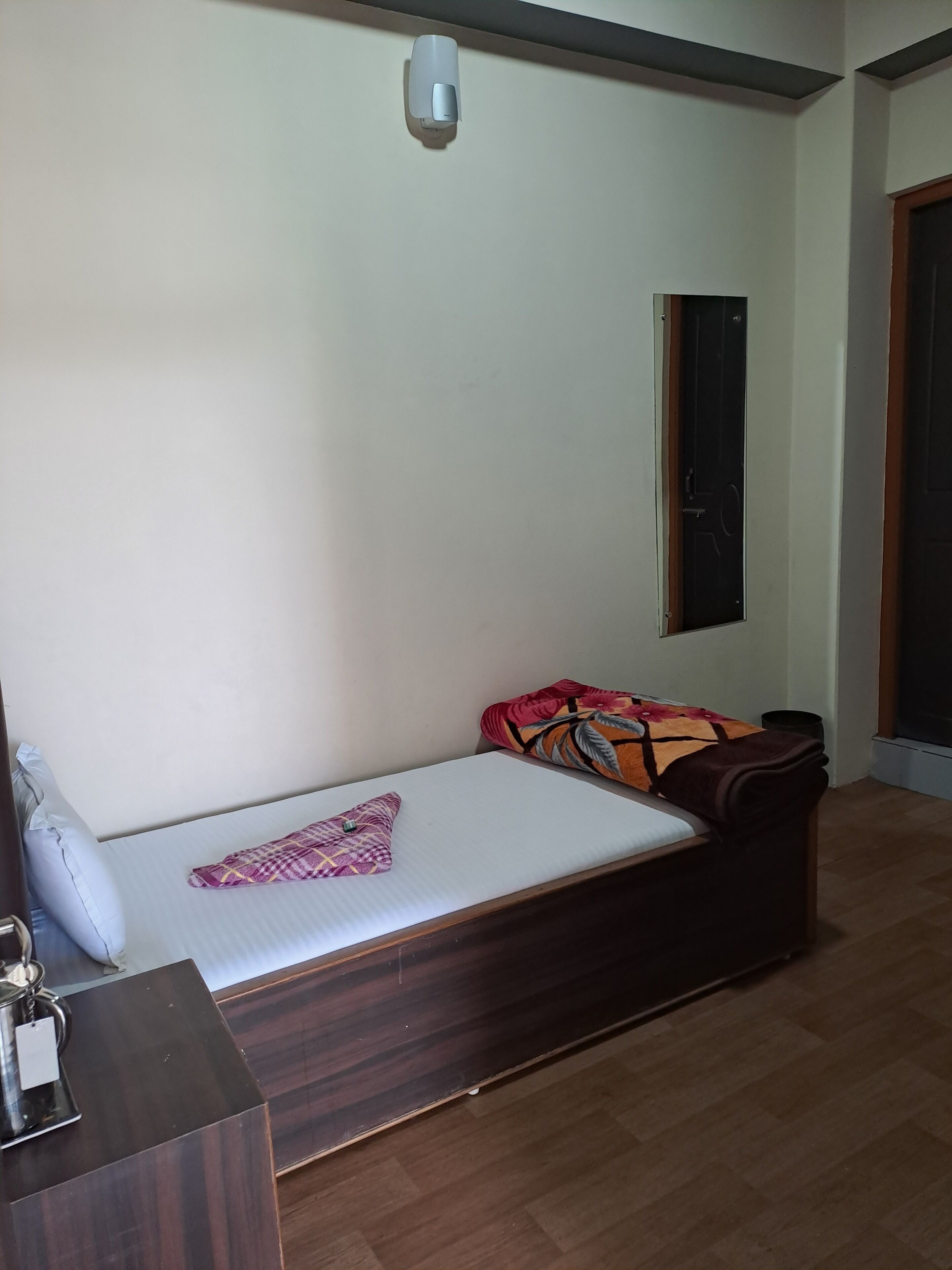 A single bed with a wooden frame, a bedside table with a kettle, and a wall-mounted light fixture.