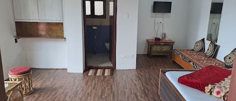Quarto com piso de madeira, cama com cobertor vermelho, televisão, espelho, mesinha com uma planta e porta que dá para um banheiro.
