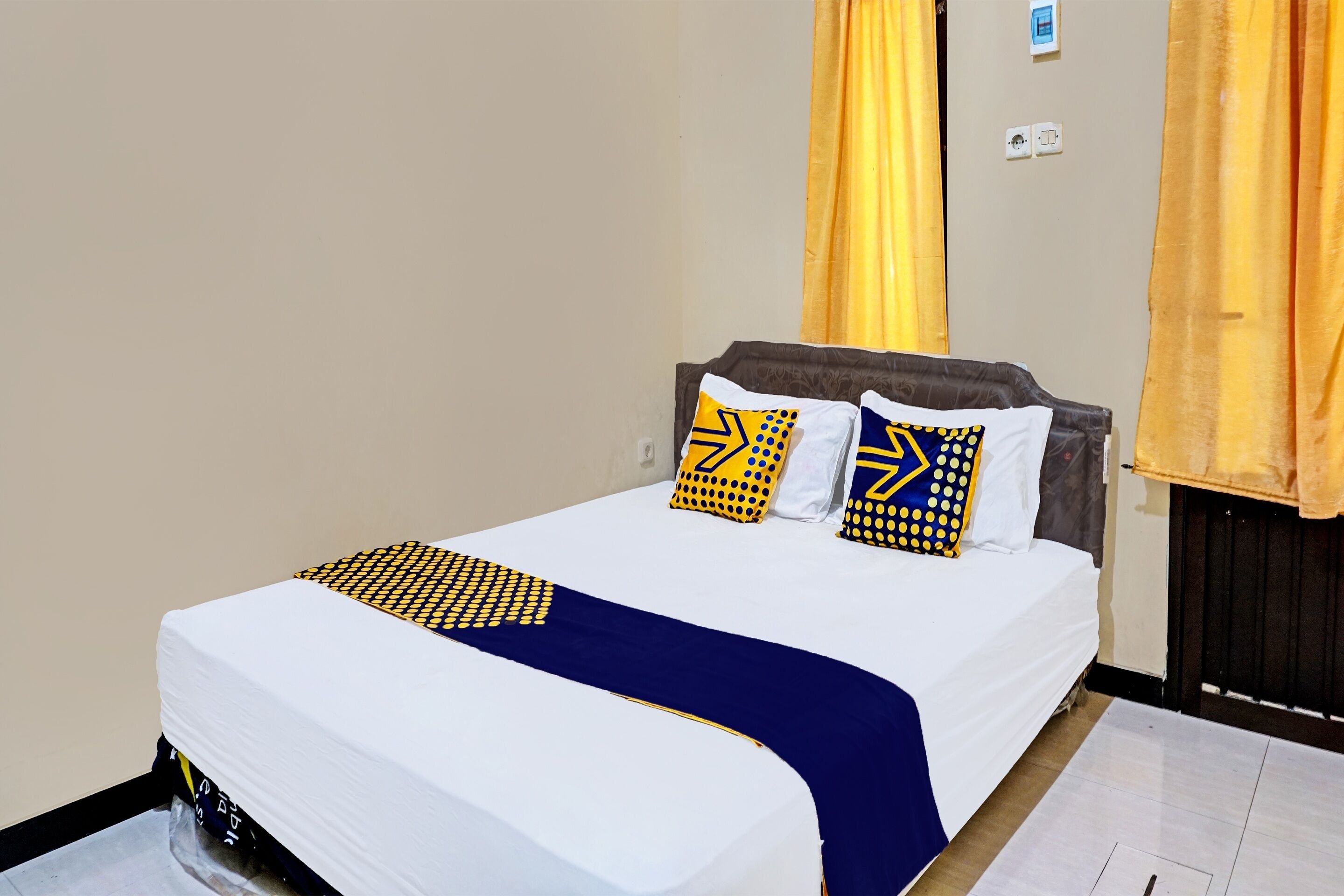 Un lit bien fait, recouvert d’une couvre-lit blanche et bleue, avec deux oreillers aux motifs jaunes et bleus, et des rideaux jaunes à la fenêtre.