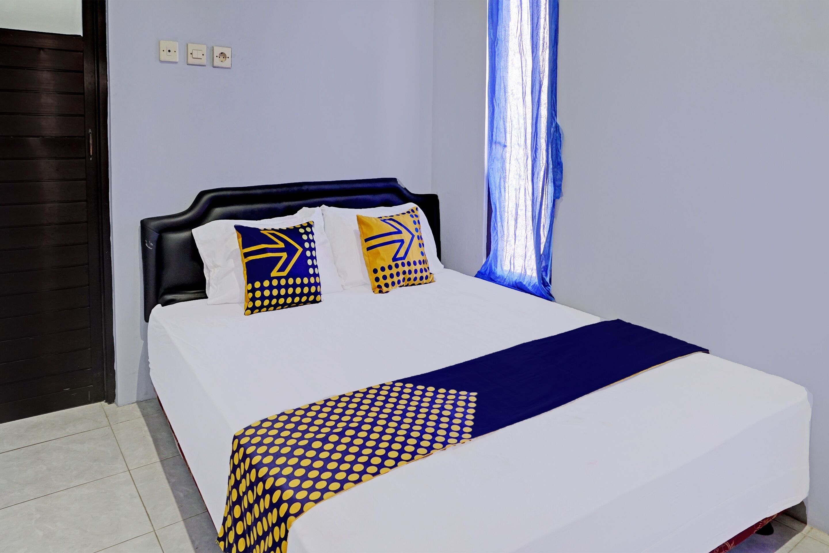Uma cama bem arrumada com um cobreleito branco e azul, duas almofadas com padrões amarelos e azuis, e uma cortina azul parcialmente aberta.