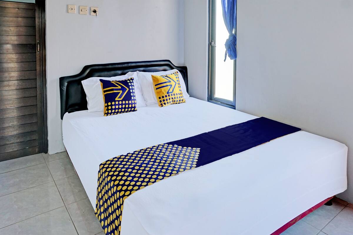 Una cama bien hecha con una colcha blanca y azul, dos almohadas con diseños amarillos y azules, y una cortina azul en la ventana.