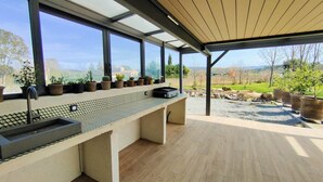 Una cucina esterna moderna con lavello integrato e barbecue, ampie finestre che offrono vista sulla campagna, e un patio in legno.