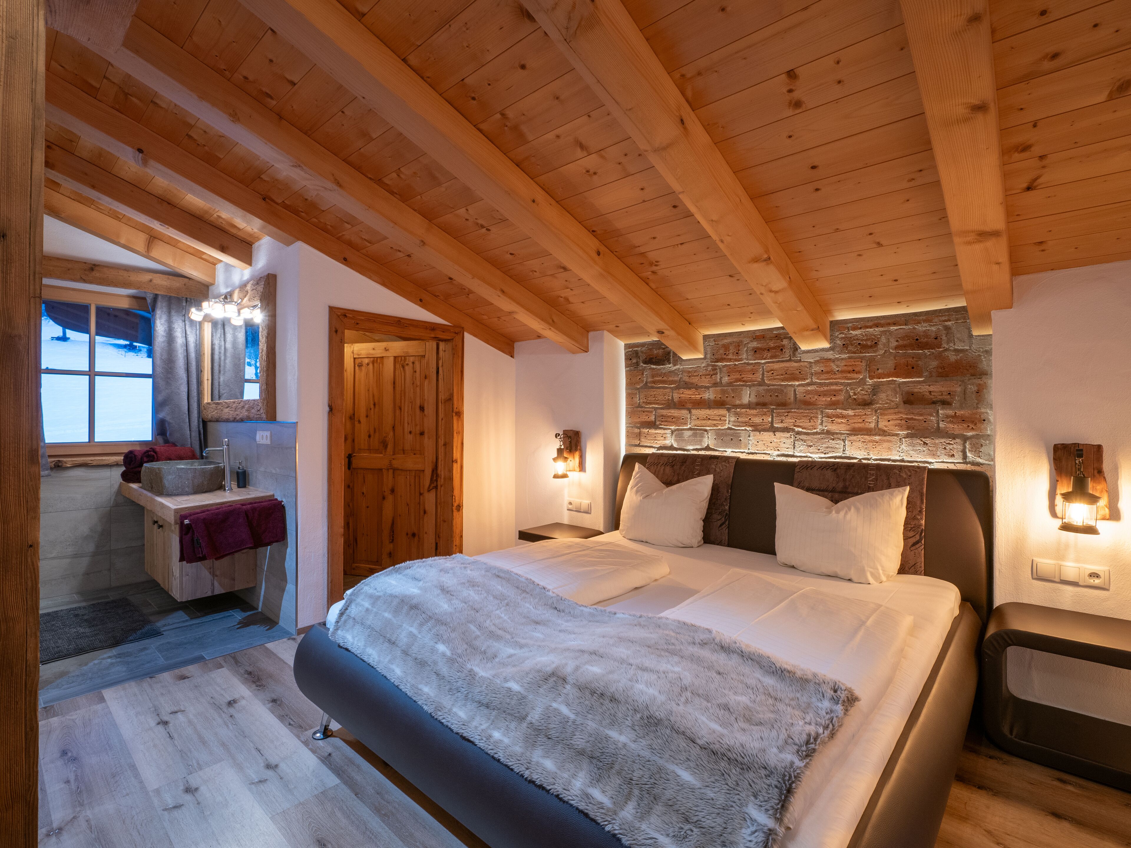 A bedroom with a wooden ceiling, a brick wall, a bed with white bedding, and a nightstand with a lamp.