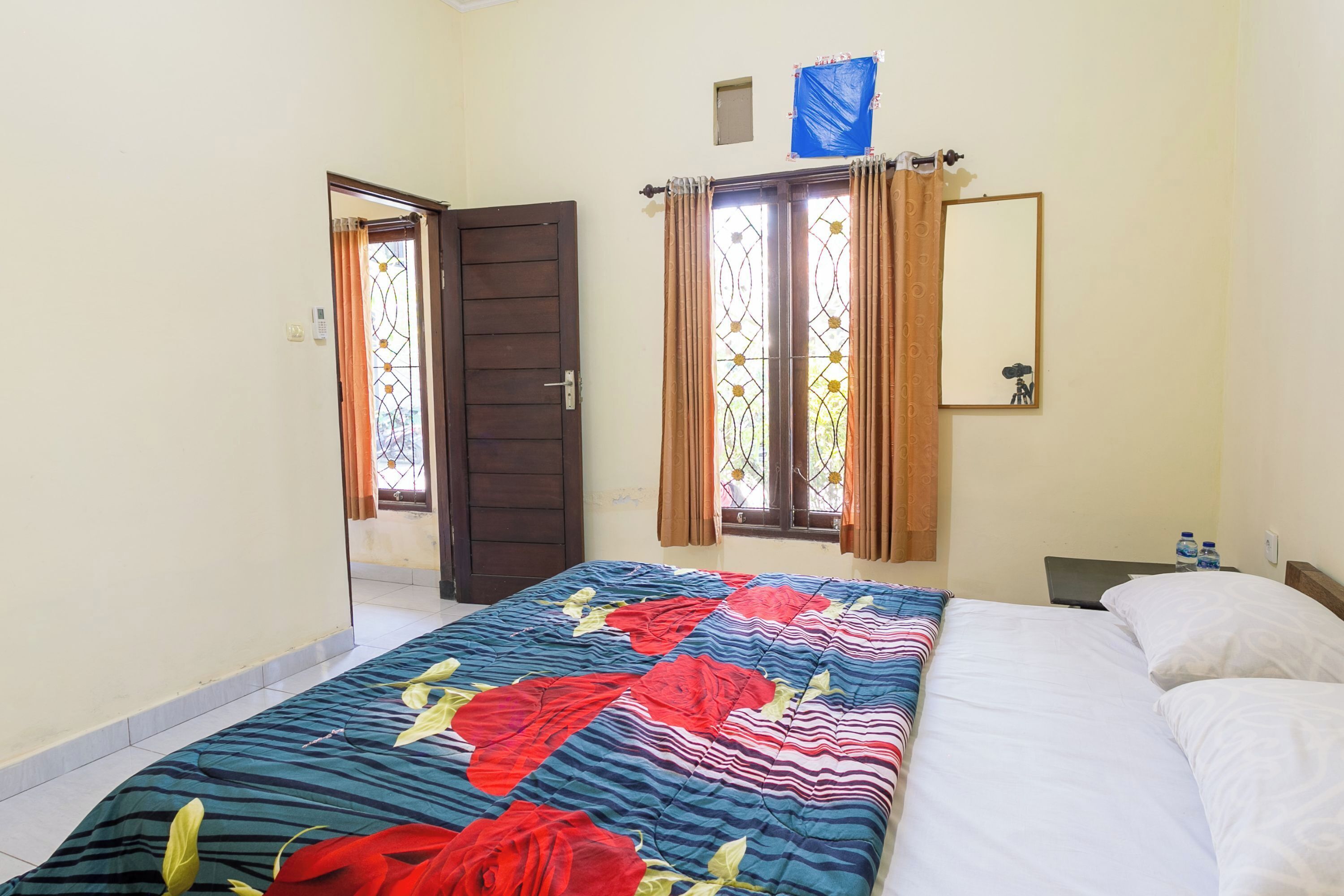 Un dormitorio con cama, dos ventanas con cortinas, una puerta, un espejo pequeño y una mesita de noche con una botella de agua.