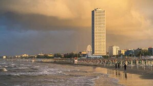 Ein Strand mit einem hohen Gebäude im Hintergrund, einer Pier mit Sonnenschirmen und Menschen, die am Ufer entlanggehen.