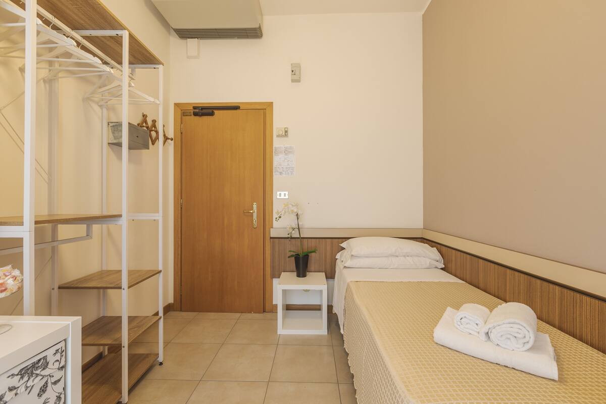 Une chambre d’hôtel avec un lit, une porte en bois, une table de chevet blanche, une étagère blanche et une plante en pot.