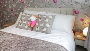 A neatly made bed with a fluffy blanket, a plush toy, and two pillows with a sheep pattern. The room has floral wallpaper and a bedside table with a lamp. - 50A Lloyd Street West (Llandudno)