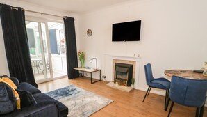 A modern living room with a sofa, a dining table with chairs, a fireplace, and a TV mounted on the wall. - 50A Lloyd Street West (Llandudno)