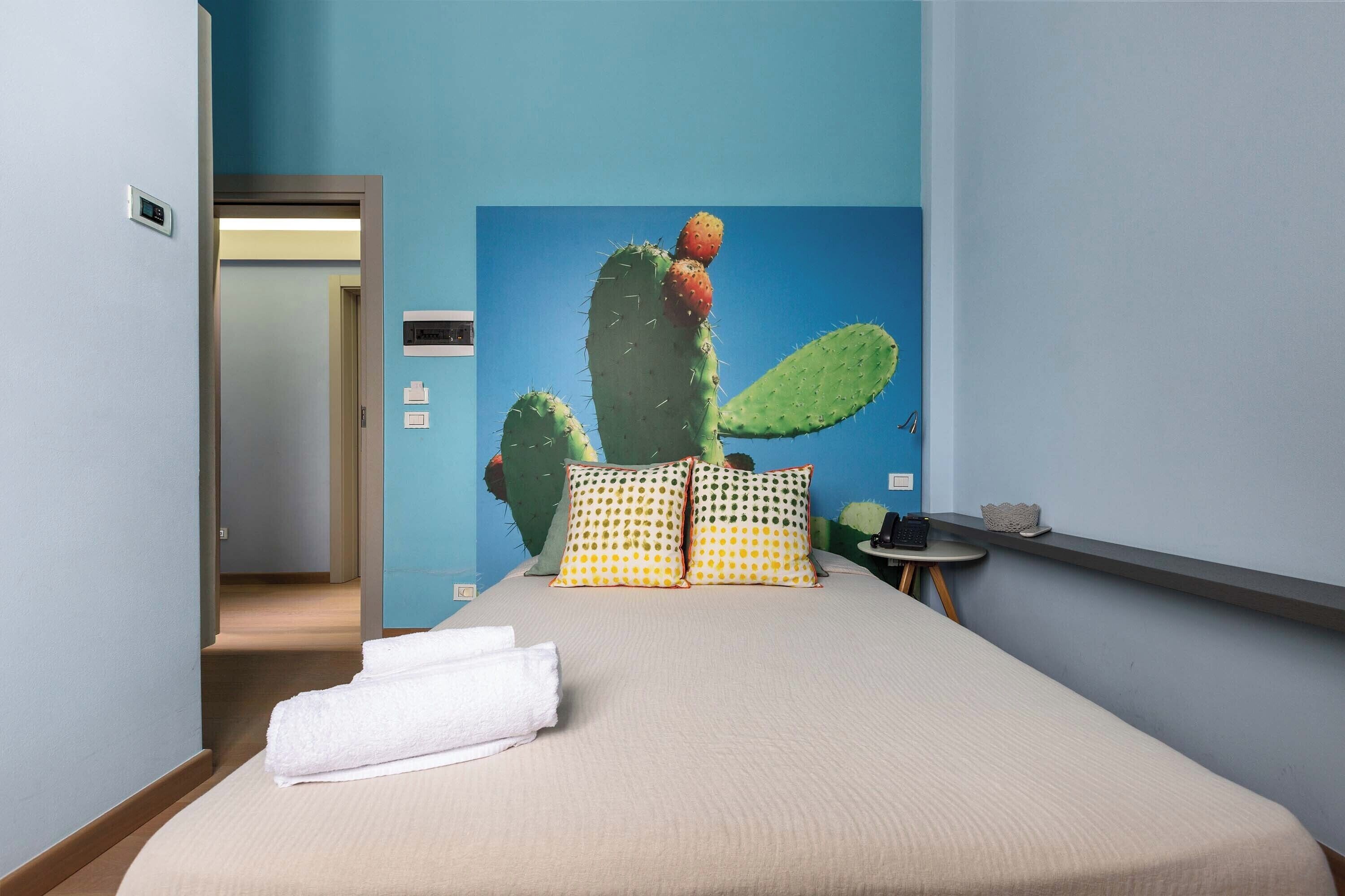 A hotel room with a bed, pillows, a wall-mounted cactus, and a small table with a cup and saucer.