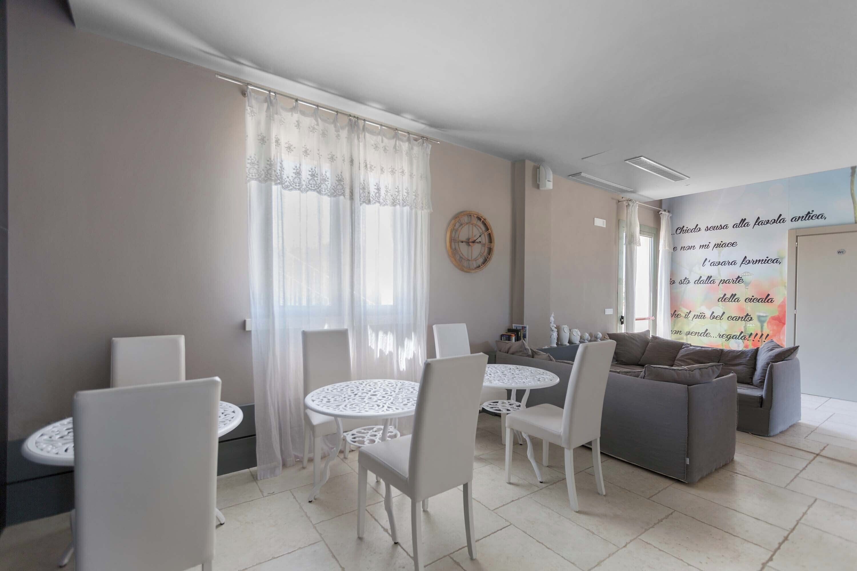 A modern living room with a grey sofa, white chairs, a round dining table, and a large window with sheer curtains.
