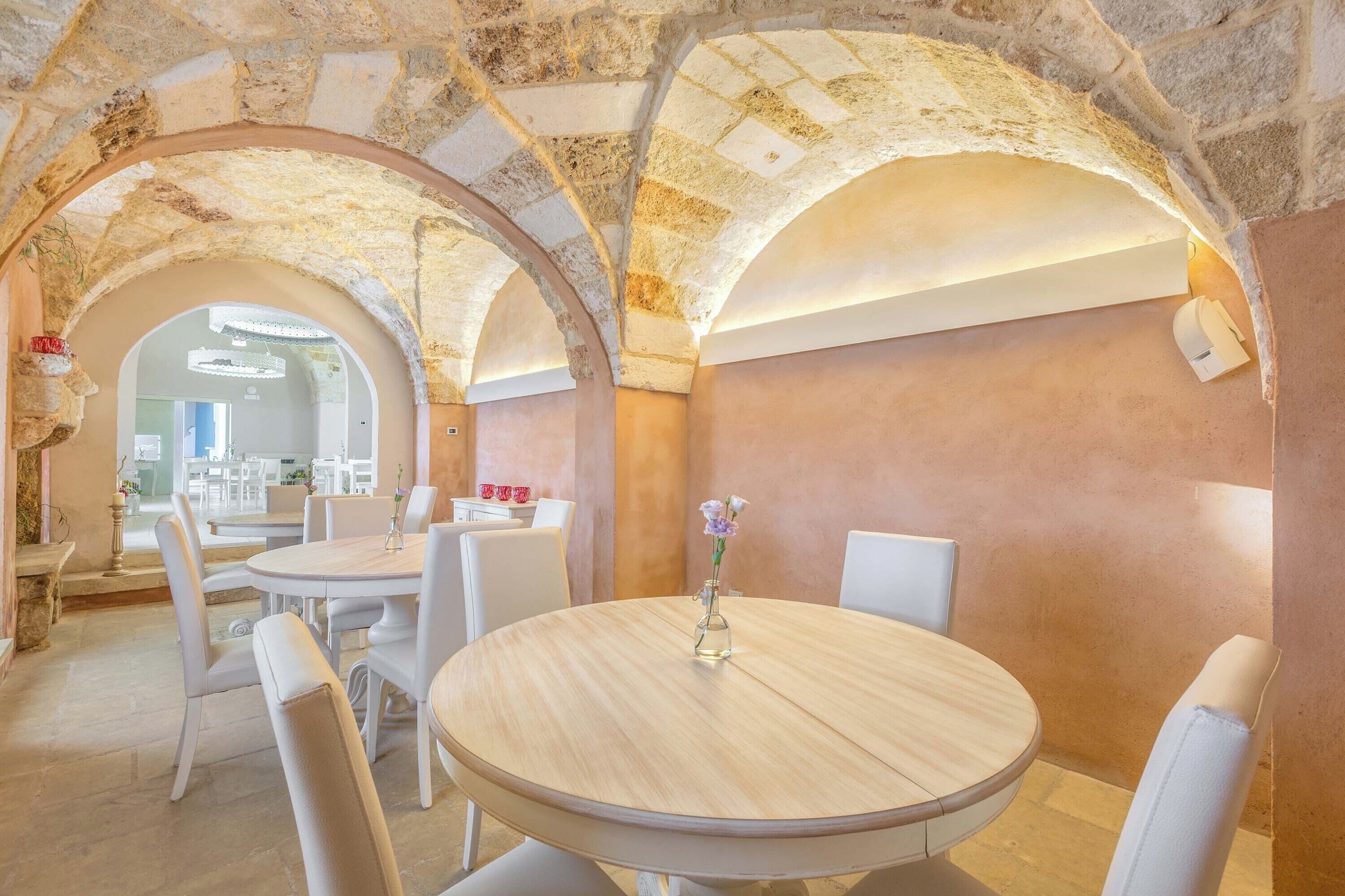 Zona pranzo con tavolo rotondo, sedie bianche e un vaso di fiori. La stanza presenta archi in pietra e una palette di colori caldi.