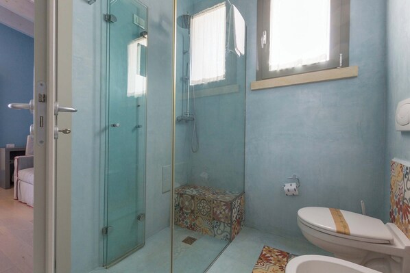Un baño con ducha de vidrio, inodoro con cubierta decorativa y alfombra estampada en el suelo.