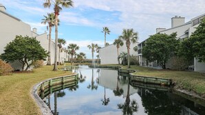 Un canal dont l’eau reflète les palmiers et les bâtiments, entouré de zones verdoyantes et sous un ciel dégagé.