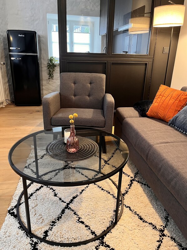 A modern living room with a glass coffee table, grey sofa, and a black refrigerator. - 2-room apartment in a historic building in Goslar's Old Town (Goslar)
